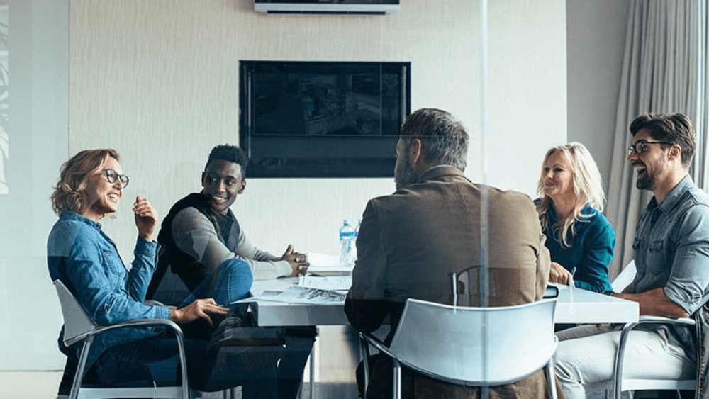 In a promotional Image for Dare to Lead Course, a group of adults gather around a conference table.